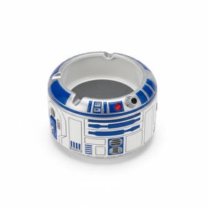 Könnte beinhalten: Ein Keramik-Aschenbecher in Form der Star Wars-Figur R2-D2. Der Aschenbecher ist weiß mit blauen und silbernen Akzenten und zeigt die ikonischen Details der Figur. Er hat ein kreisförmiges Design mit grauem Innenraum.