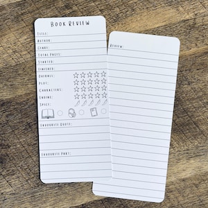 May include: Two white rectangular paper bookmarks with black printed text and lines. The left bookmark features fields for book title, author, genre, pages, dates, star ratings, chilli pepper spice levels, and icons. The right bookmark has a review section.