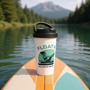 Könnte beinhalten: Weißer Reisebecher mit schwarzem Deckel und Griff, mit dem Wort "FLOATS" in Grün und einer türkisfarbenen Grafik einer Person, die Yoga macht. Der Becher steht auf einem Paddleboard mit einem Berg und einem See im Hintergrund.