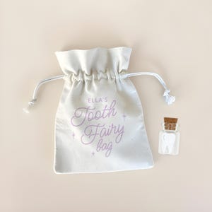May include: A white drawstring bag with the text "Ella's Tooth Fairy bag" in lavender script. A small glass jar with a cork stopper and a tooth-shaped object is next to the bag. The background is a solid, light beige color.