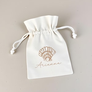 May include: A white cotton drawstring bag with a brown seashell design and the name "Arianna" printed on the front.