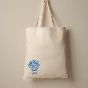 May include: Cream-colored tote bag with a blue seashell graphic and the word "April" printed below it. The bag has two cream-colored handles and is made of a sturdy fabric, suitable for carrying groceries or other items.