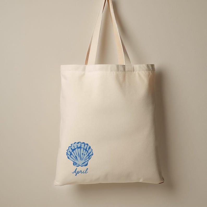 May include: Cream-colored tote bag with a blue seashell graphic and the word "April" printed below it. The bag has two cream-colored handles and is made of a sturdy fabric, suitable for carrying groceries or other items.