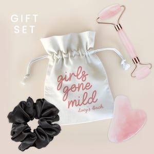 May include: A gift set featuring a white drawstring bag with the text "girls gone mild" and "Lucy's Bach" in pink. The set includes a black scrunchie, a pink rose quartz face roller, and a pink rose quartz gua sha tool.