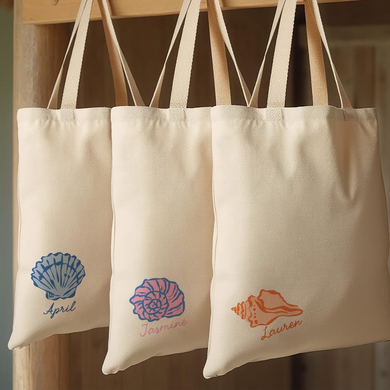May include: Three cream-colored tote bags with seashell designs. Each bag features a different seashell graphic in blue, pink, and orange, with a name printed below each shell. The bags are hanging on a wooden rack.