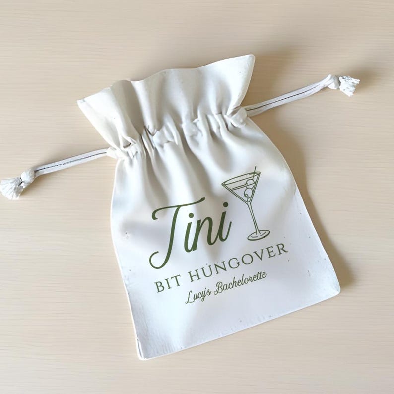 May include: White cotton drawstring bag with the text "Tini Bit Hungover Lucy's Bachelorette" and a green line drawing of a martini glass with an olive.