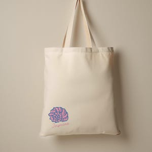 May include: Cream-colored tote bag with a seashell design in pink and blue. The word "Jasmine" is written in cursive below the seashell. The bag has matching cream-colored handles.