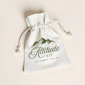 May include: A cream-colored drawstring bag with a green mountain graphic and the words "Altitude KIT" in a script font. The text "AVA + CHARLIE | VAIL, CO" is printed below. The bag is tied with a white drawstring.