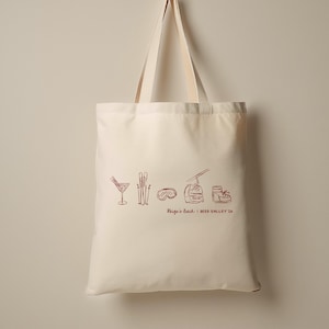 May include: Cream-colored tote bag with a maroon line drawing of a cocktail, skis, goggles, a ski lift, and a ski boot. The text "Paige's Bach | Deer Valley 24" is below the drawings. The bag has two cream-colored handles.