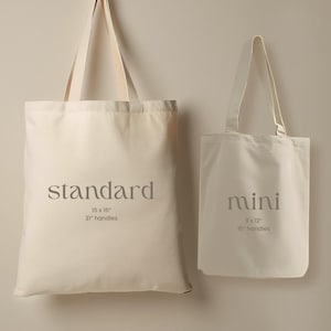 May include: Two off-white canvas tote bags. The larger bag is labeled "standard" and measures 15 x 16 inches with 21-inch handles. The smaller bag is labeled "mini" and measures 9 x 12 inches with 15-inch handles. Both bags have long handles.