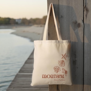 May include: A white canvas tote bag with a brown design of seashells and a starfish. The bag has the text "Montauk" in brown letters and "Anna & Matthew" in smaller brown letters below.