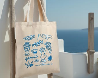 May include: A white canvas tote bag with blue illustrations of Greek food and culture, including fish, grapes, olives, a pitcher, a cup of coffee, and a bust of a woman. The text "Yamas" is printed in blue on the bag, along with the words "Cali & Andrea - Paros '23".