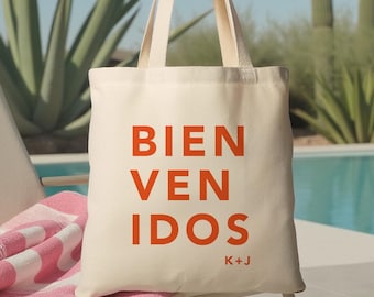 May include: Cream-colored tote bag with the words "BIEN VENIDOS" in orange block letters. The bag has long handles and the initials "K+J" below the text. A pink and white striped towel rests on a chair next to the bag.