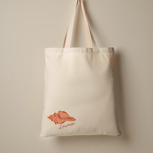 May include: Cream-colored tote bag with matching handles. A coral-colored seashell graphic and the name "Lauren" are printed on the front. The bag is hanging against a neutral background.