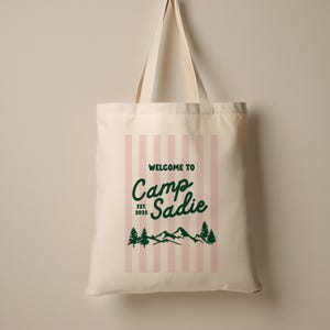 May include: Cream-colored tote bag with pink vertical stripes and the text "WELCOME TO Camp Sadie EST. 2025" in green. The design includes mountain and tree graphics.