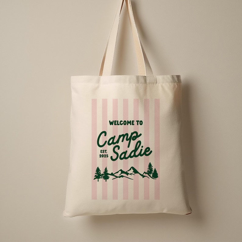 May include: Cream-colored tote bag with pink vertical stripes and the text "WELCOME TO Camp Sadie EST. 2025" in green. The design includes mountain and tree graphics.