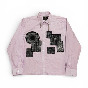 May include: A pink and white striped button-down shirt with black, textured geometric patches. A silver chain connects the collar buttons. The long-sleeved shirt has buttoned cuffs. The shirt is a unique design.