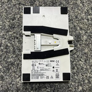 Puede incluir: Un componente electrónico blanco con almohadillas y detalles en negro. El componente tiene un soporte metálico y varias etiquetas con texto y símbolos. El componente es rectangular con esquinas redondeadas.