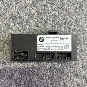 Könnte beinhalten: Ein schwarzes elektronisches Steuermodul mit mehreren Anschlüssen. Das Modul trägt unter anderem die Aufschrift "HKFM 21 High DSL", "brose" und ein BMW-Logo. Das Modul ist rechteckig und hat Anschlüsse.