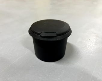 Op de afbeelding: Een zwarte, cilindervormige container met een bijpassend deksel. Het deksel heeft een verzonken gedeelte voor gemakkelijk openen. De container is waarschijnlijk gemaakt van plastic of een vergelijkbaar materiaal. Het ontwerp is eenvoudig en functioneel.