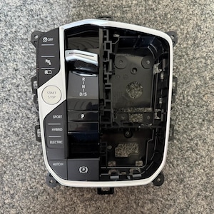 Op de afbeelding: Zwart en zilver auto-console met knoppen voor verschillende functies, waaronder "OFF", "START STOP", "SPORT", "HYBRID" en "ELECTRIC". De console heeft ook een versnellingsindicator met de opties "R", "N", "D/S" en "P".