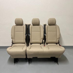 Könnte beinhalten: Drei beigefarbene Fahrzeugsitze mit Kopfstützen und integrierten Sicherheitsgurten. Die Sitze sind nebeneinander angeordnet, was auf eine Bankkonfiguration hindeutet. Die Sitze bestehen aus einem glatten, lederartigen Material und sind auf einem schwarzen Metallrahmen montiert.