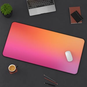 May include: A rectangular desk mat with a pink to orange gradient design. A white computer mouse sits on the mat. Other items include a laptop, a small potted plant, a notebook, and writing utensils.