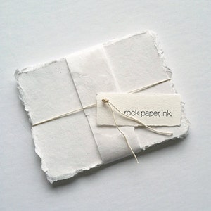 May include: A stack of white handmade paper with rough edges, tied with twine and a tag that reads "rock paper, ink".