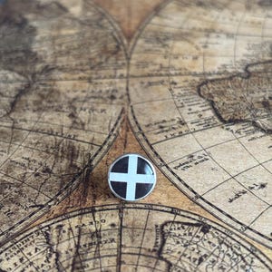 May include: A circular pin with a black and white cross design, set within a silver-toned frame. The pin is displayed on a vintage map, giving it a classic, historical feel. The design is simple and striking, suitable for various uses.