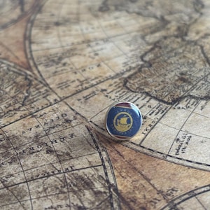 May include: A circular navy blue and gold enamel pin featuring a ship design. The pin is set against a vintage world map background, evoking a travel or nautical theme. The pin's diameter is approximately 2 cm.