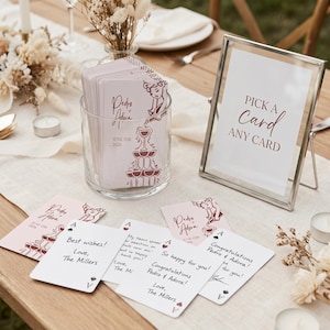 Peut inclure: Une table de réception de mariage avec un bocal en verre rempli de cartes roses, un panneau encadré avec l'inscription "PICK A CARD ANY CARD" et plusieurs cartes avec des messages manuscrits. Les cartes présentent une illustration de tour de champagne.