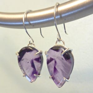 May include: A pair of silver dangle earrings with a teardrop-shaped amethyst stone set in a silver bezel. The amethyst stones are a light purple color and have a slightly cloudy appearance.