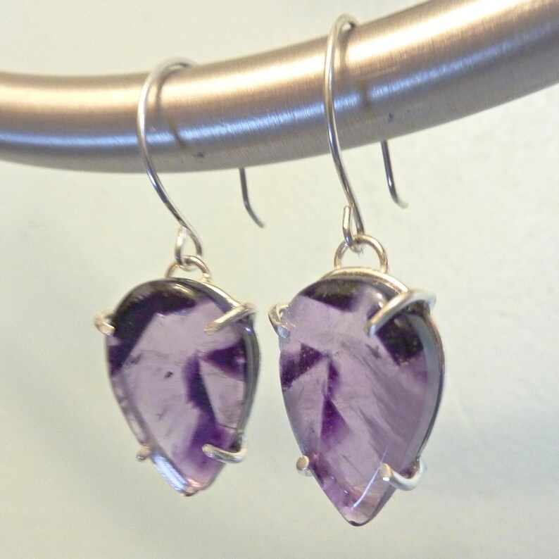 May include: A pair of silver dangle earrings with a teardrop-shaped amethyst stone set in a silver bezel. The amethyst stones are a light purple color and have a slightly cloudy appearance.