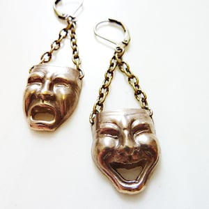 Op de afbeelding: Een paar zilveren oorbellen met een ketting en een hanger in de vorm van een masker. Het ene masker is een lachend gezicht en het andere is een huilend gezicht.