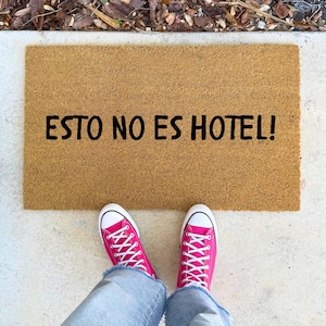 May include: A rectangular doormat made of natural fibers with the text "ESTO NO ES HOTEL!" in black. The mat is on a concrete surface, with a person wearing pink sneakers and blue jeans.