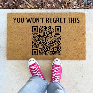 May include: A brown doormat with the text "You won't regret this" and a black and white QR code. A pair of pink sneakers are standing on the doormat.
