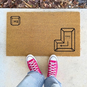 May include: A brown doormat with the words "ESC" and "Enter" printed on it. The "Enter" key is depicted as a stylized arrow pointing to the left. Two pink sneakers are standing on the concrete floor in front of the doormat.