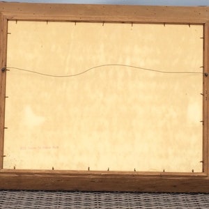 May include: The back of a wooden picture frame with a light brown finish. The frame is hanging on a wall. The back of the picture is visible, and it is a light beige color. There is a small piece of paper with the text "Paul Gauguin, "La Siesta" printed on it.