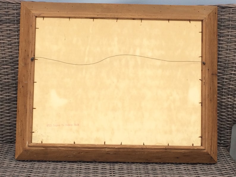 May include: The back of a wooden picture frame with a light brown finish. The frame is hanging on a wall. The back of the picture is visible, and it is a light beige color. There is a small piece of paper with the text "Paul Gauguin, "La Siesta" printed on it.