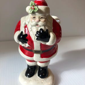 May include: A ceramic Santa Claus figurine, featuring a red hat and suit with white trim, black gloves and boots. The figurine has a white beard and a sprig of holly on the hat. The base is white and snow-like.