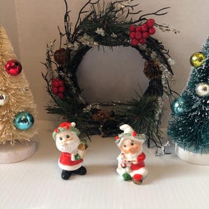 May include: Two small ceramic Christmas figurines, one depicting Santa Claus holding a gift and the other a gnome holding a small tree. Both figurines are wearing red and white outfits with green accents. The figurines are positioned in front of a Christmas wreath with red berries and pine cones.