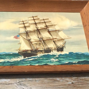 May include: A framed painting of a large sailing ship with white sails and a blue and red American flag. The ship is sailing on a blue ocean with white waves.