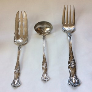 May include: Three silver serving pieces with ornate grapevine designs. The pieces include a serving fork, a serving spoon, and a serving fork with a decorative cut-out design.