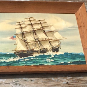 May include: A framed painting of a large sailing ship with white sails, a dark hull, and an American flag flying from the mast. The ship is sailing on a blue and green ocean with white waves.