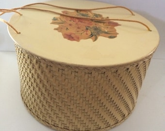 May include: A round, woven wicker basket with a cream-colored lid. The lid has a floral design in shades of orange, yellow, and brown. The basket has two rope handles.
