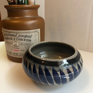 May include: A ceramic bowl with a blue and white leaf pattern. The bowl has a dark blue rim and a gray interior. A brown jar with paintbrushes and the text "fashioned mustard" is in the background.