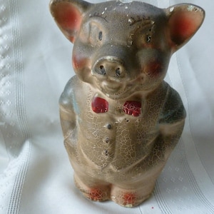 May include: A vintage ceramic piggy bank in the shape of a pig wearing a blue and white shirt and a red bow tie. The piggy bank has a cracked and weathered surface.