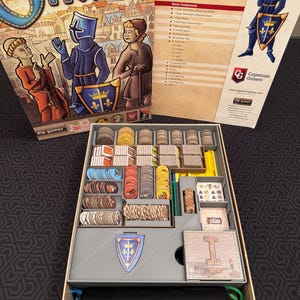 May include: The board game "Orléans" is displayed open, revealing game components. The box art features medieval figures and the game's title. Inside, various tokens, tiles, and game pieces are organized in a tray, ready for play.