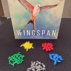 May include: A board game box with the title "Wingspan" and a watercolor illustration of a bird with outstretched wings. Various game pieces, including yellow, green, red, gray, and blue components, are scattered in front of the box.