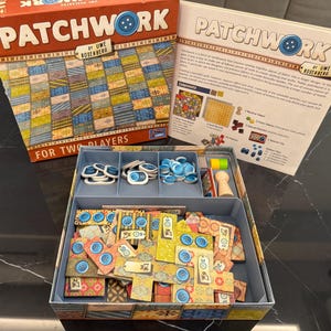 Puede incluir: El juego de mesa "Patchwork" de Uwe Rosenberg se muestra abierto, revelando piezas del juego e instrucciones. La caja es roja con un diseño de colcha de retazos. El juego incluye fichas de botones, tableros y piezas de tela. El juego es para dos jugadores.